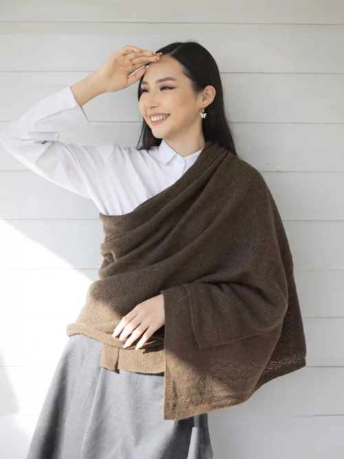 Femme souriante portant un châle yack marron chocolat, habillée d'une chemise blanche et d'une jupe grise, posant devant un mur en bois. Ce châle en laine 100 % mongole est parfait pour se réchauffer avec style en hiver.