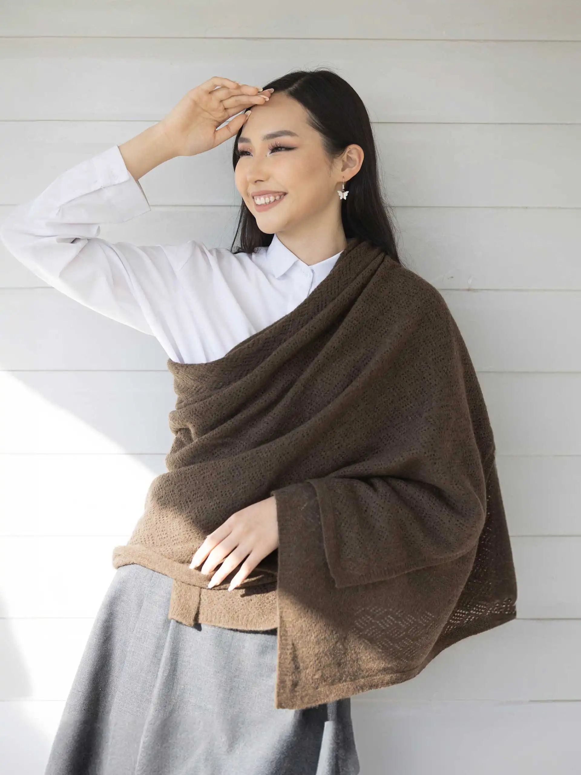 Femme souriante portant un châle yack marron chocolat, habillée d'une chemise blanche et d'une jupe grise, posant devant un mur en bois. Ce châle en laine 100 % mongole est parfait pour se réchauffer avec style en hiver.