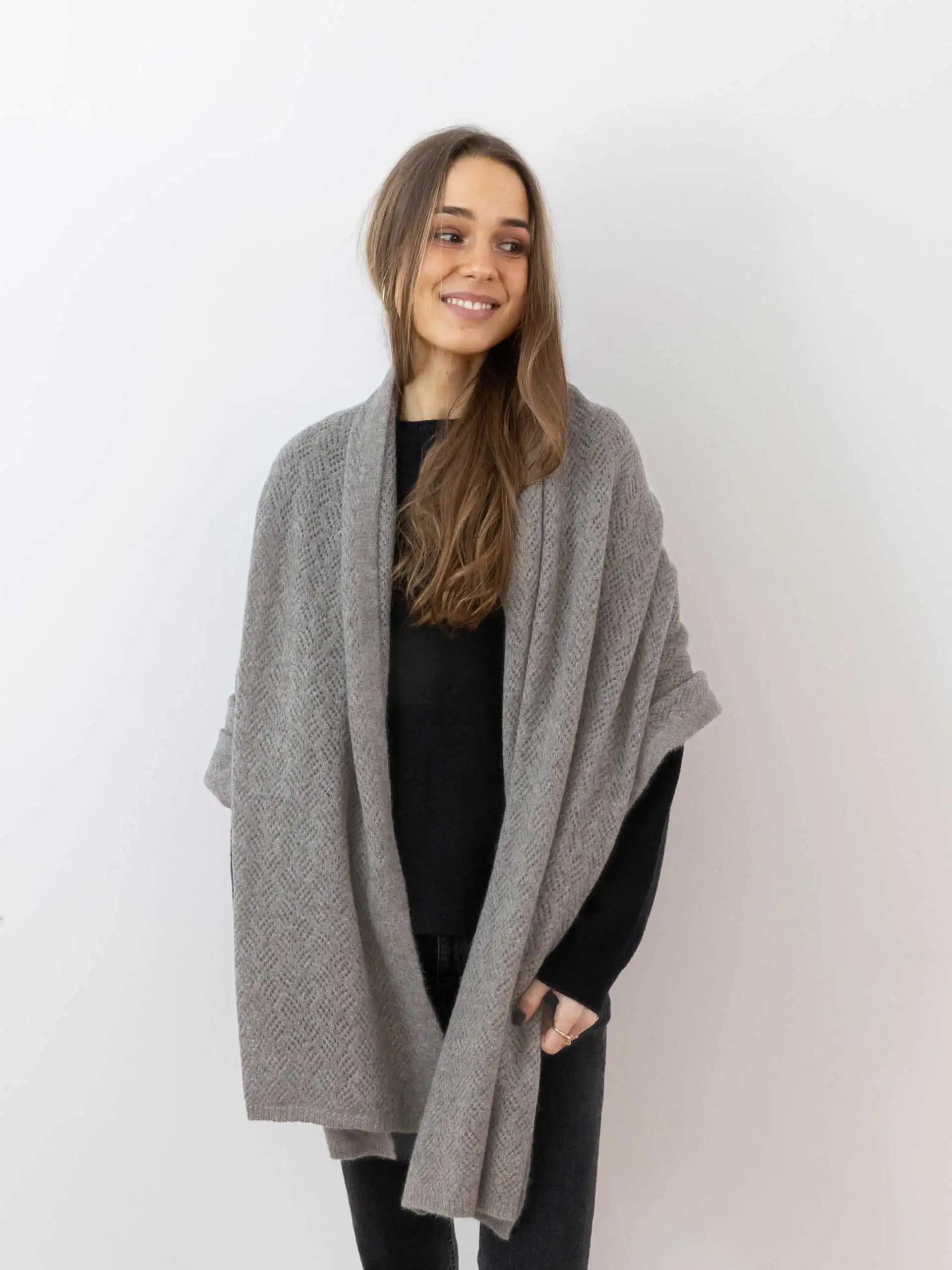 Femme souriante portant une étole yack gris cendré, habillée d'un pull noir et posant devant un fond blanc. Cette étole en laine 100 % mongole est parfaite pour se réchauffer avec style en hiver.