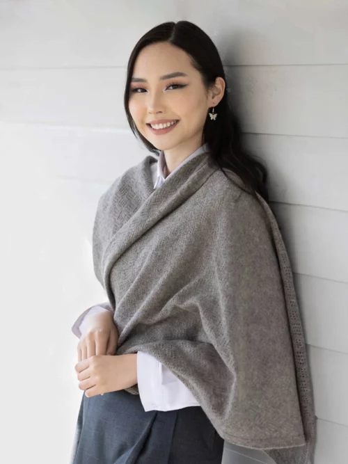 Femme portant une étole yack gris cendré, habillée d'une chemise blanche et d'une jupe grise, posant devant un mur en bois. Cette étole en laine 100 % mongole est parfaite pour se réchauffer avec style en hiver.