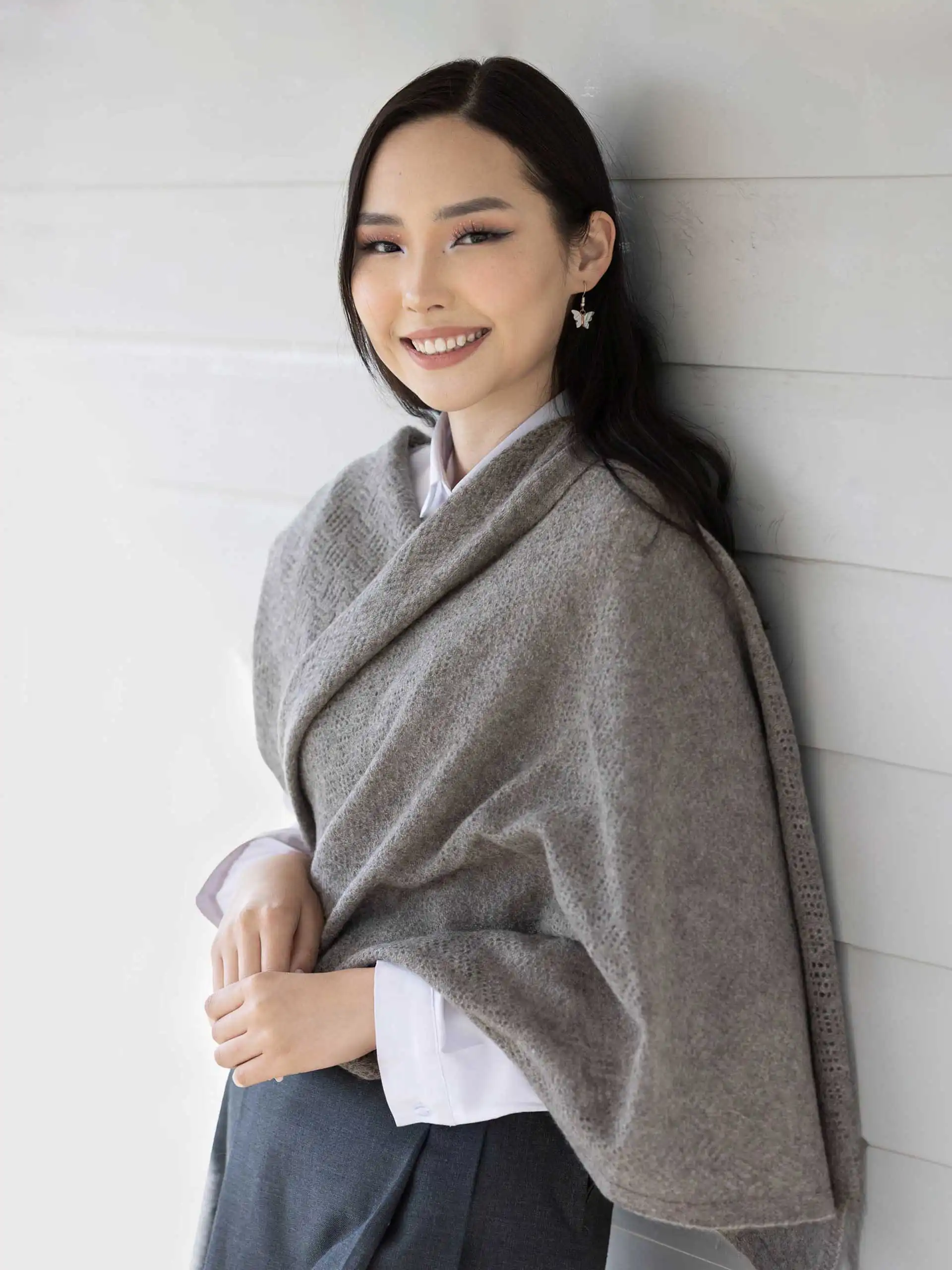 Femme portant une étole yack gris cendré, habillée d'une chemise blanche et d'une jupe grise, posant devant un mur en bois. Cette étole en laine 100 % mongole est parfaite pour se réchauffer avec style en hiver.