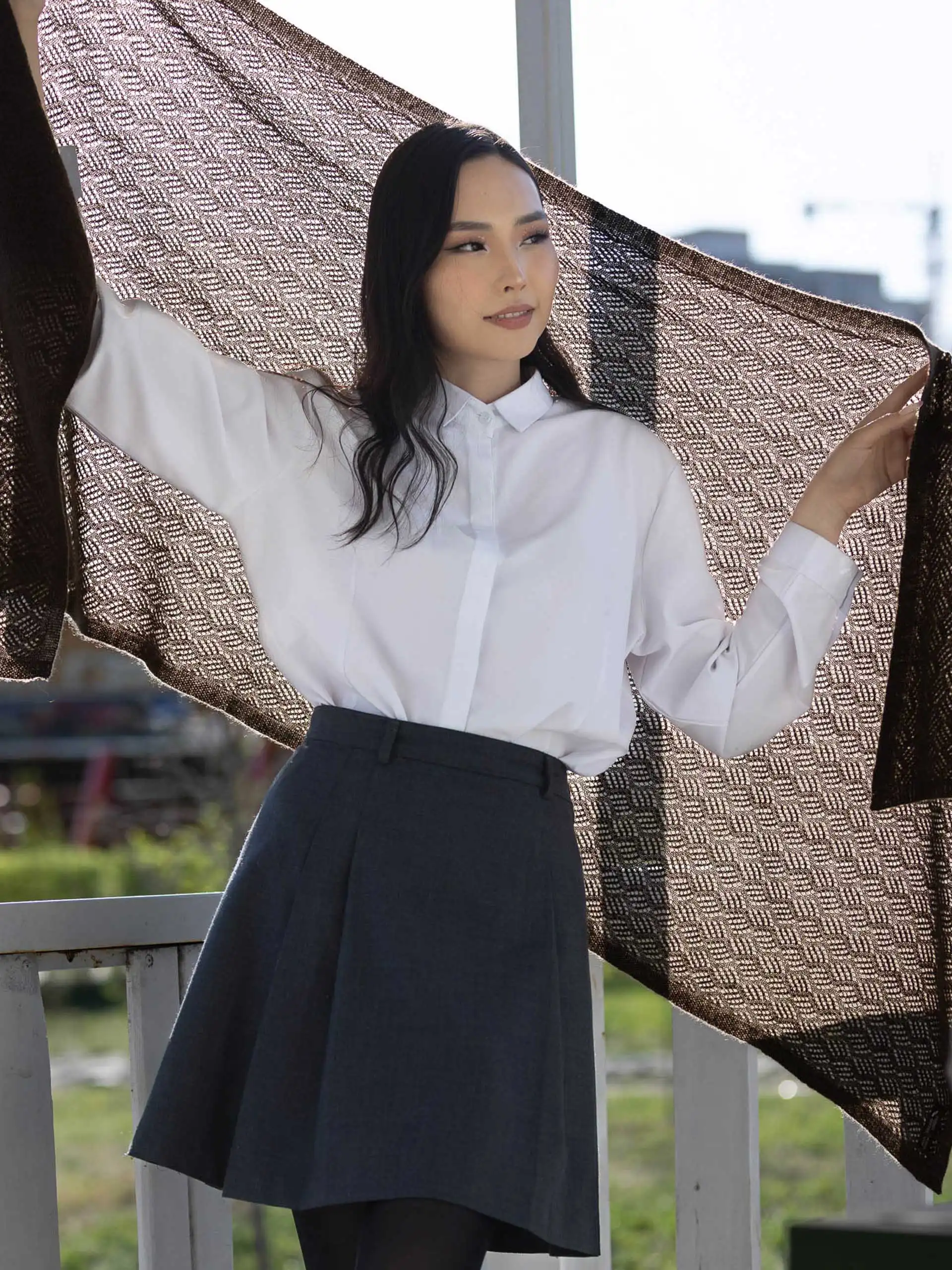 Femme portant une étole yack marron foncé point pointelle, vêtue d'une chemise blanche et d'une jupe grise, tenant l'étole déployée pour montrer la finesse du tricot. Un accessoire élégant pour les saisons fraîches.
