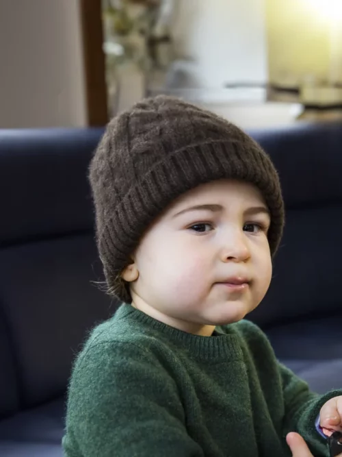 Enfant assis sur un canapé portant un bonnet yack marron foncé torsadé de Mongolie, avec une expression sérieuse.
